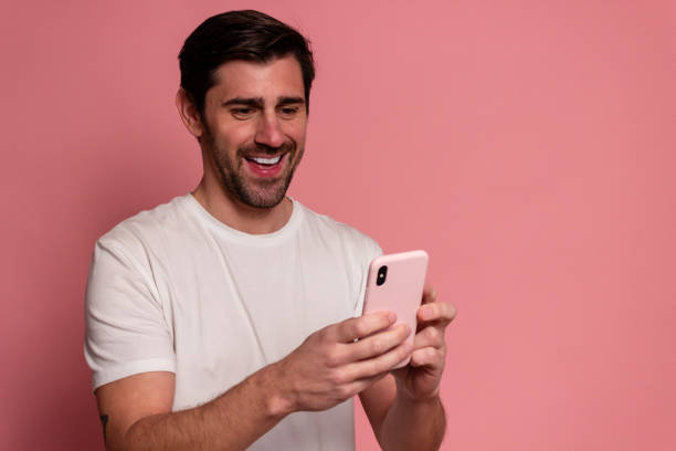 istock Cheerful young man in white t-shirt is using mobile phone and smiling while standing against pin 1895700835