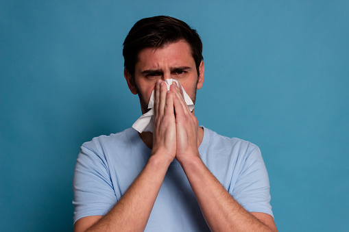 Portrait of a sick man sneezing into tissue on blue background
