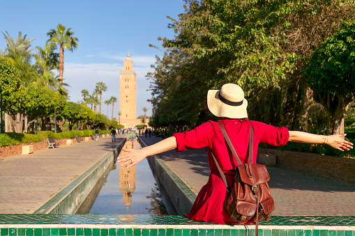 Young woman dressing in red with open arms looking the mosque of Koutoubia in Marrakesh