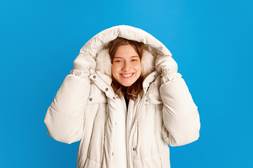 Portrait of adorable happy young girl, student in warm winter outfit smiling looking at camera against blue studio background. Concept of New Year, Christmas, winter holidays, tourism, vacation.
