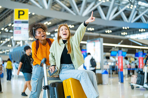 Happy woman friends holding passport and suitcase walking together to airline check in counter in airport terminal. Attractive girl enjoy and fun travel on holiday vacation with airplane transportation