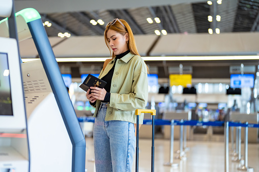 Asian woman holding passport using self Check-in kiosk machine getting airline ticket boarding pass at airport terminal. People travel on holiday vacation and global airplane transportation concept.