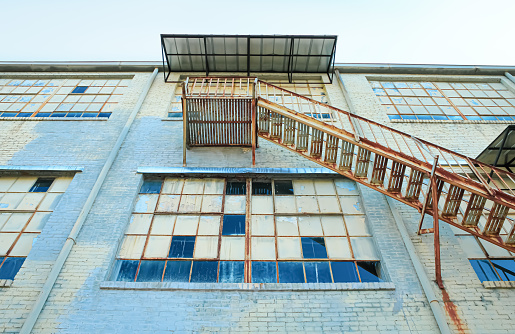 Looking up at a rusted fire escape on the side of an old factory building with blue and green colors