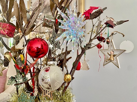 Horizontal close up of part of christmas tree with decorations of glass balls, handmade dolls and more on old man banksia tree branches agsainst white wall in domestic Australian home