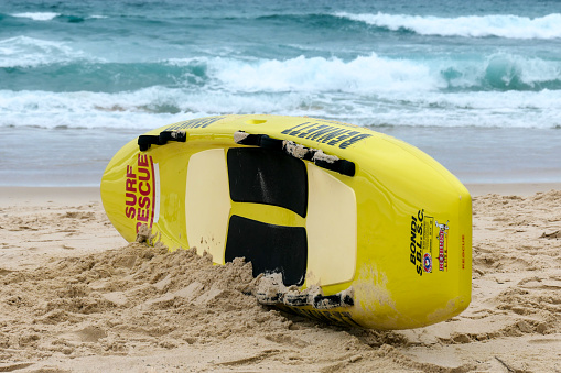A surfboard of the volunteer Surf Lifesavers placed upright and ready to be used at Bondi Beach, Sydney.  Despite the holiday season, the beach is quite empty because of the overcast conditions.  This image was taken on an overcast afternoon on 31 December 2023.