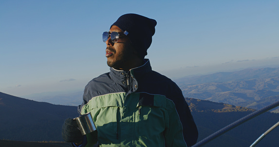 African American man in sunglasses stands near tent and looks at beautiful forest valley. Tourist drinks cup of hot tea and warms up on top of mountain hill. Backpacker stopped to rest during a hike.