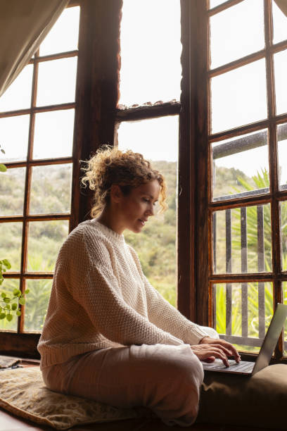 Woman sit down on the floor an duse laptop in cozy wooden cabin with nature outdoors view. Concept of alternative healthy and technology lifestyle and small business. People using notebook indoor la mujer se sienta en el suelo con una computadora portátil duse en una acogedora cabaña de madera con vista al aire libre de la naturaleza. concepto de estilo de vida alternativo, saludable y tecnológico y pequeña empresa. personas que usan notebook e