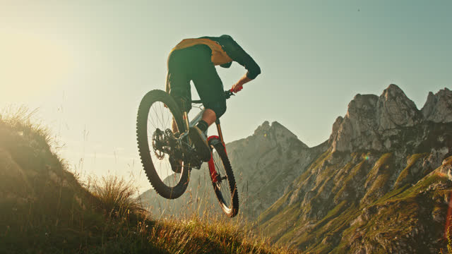 SUPER SLOW MOTION TIME WARP EFFECT Male Mountain Biker Jumping with Bicycle Over Grassy Hill against Idyllic Rocky Mountains during Sunny Day
