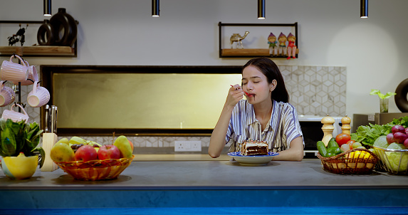 Happy Indian alone teenager girl sitting in modern kitchen eating  piece of cake at indoor house. Overjoyed beautiful cute teen age female celebrate birthday having fun enjoying holiday inside home