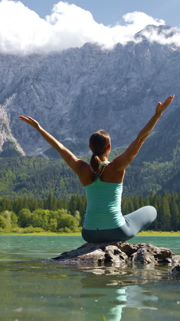 Rear View of Female Hiker Practicing Yoga Sitting on Rock in Lake amidst Rocky Mountains. Laghi di Fusine.