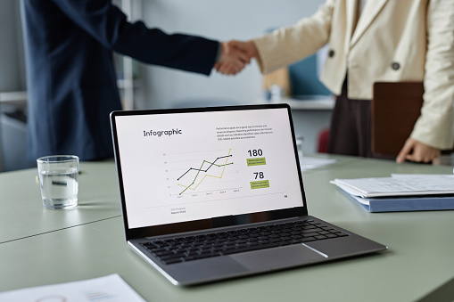 Close up shot of operating laptop with infographic on screen, unrecognizable business partners shaking hands in blurred background