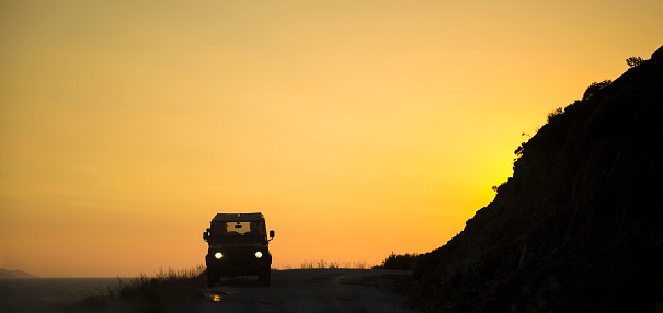 Silhouette off-road car on the road between mountains and sea during a sunset.