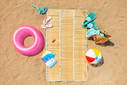 Beach accessories around straw cover on the sand on the beach. Magazines, water ball, beach bag, tower, sunscreen, water sunscreen, sea bed, beach slippers, diving goggles, diving fins