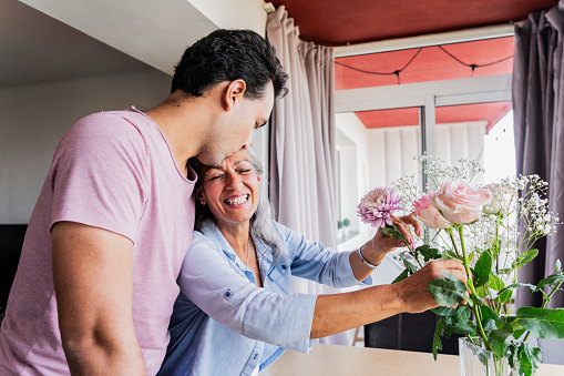 Young man kissing his mother's forehead while she takes care of bouquet rose at home