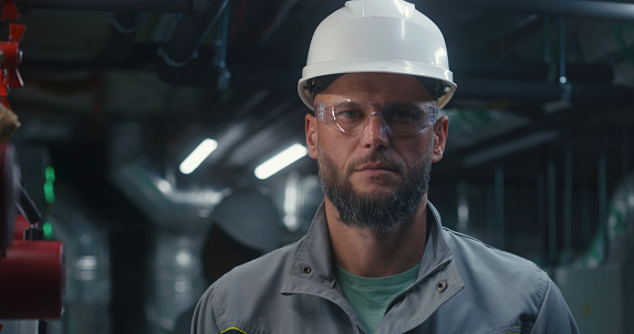 Professional Caucasian engineer stands and looks at camera. Industrial specialist or worker in safety uniform, goggles and hard hat works on modern manufacturing factory, plant or energy facility.