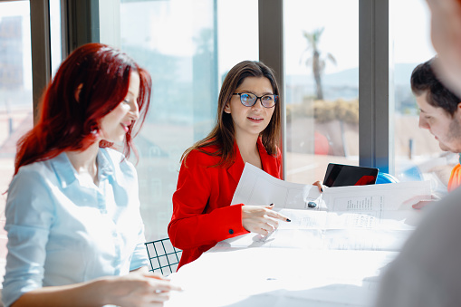 Step into the dynamic world of creativity with this captivating stock photo, featuring a stylish businesswoman in a red jacket and glasses leading a meeting in a creative or architectural office. Two young creative female colleagues and a male worker join the meeting. The positive and efficient atmosphere is evident as everyone is smiling and enjoying the collaborative session. The meeting takes place in a modern office's conference room, bathed in sunlight through large windows. Ideal for conveying creativity, teamwork, and positive work culture, this photo is perfect for business-related promotions, creative industry publications, and social media content celebrating successful collaboration. Stylish businesswoman, Creative meeting, Modern office, Architectural office, Red jacket, Glasses, Young creative colleagues, Male worker, Positive, Efficient, Smiling, Sunlight, Large windows, Stock photo, iStock, Business-related promotions, Creative industry publications, Social media content, Successful collaboration, Teamwork, Positive work culture