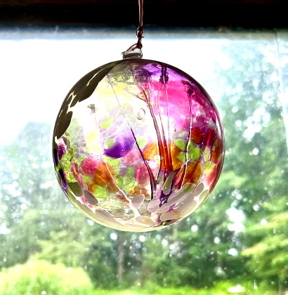 A unique large glass globe with multiple designs and colours hangs by a window.