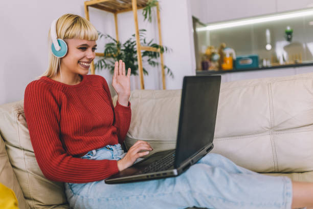 istock Smiling woman using laptop for a video call while sitting on couch at home.  Woman using laptop and having an online meeting while working from home 1897422050