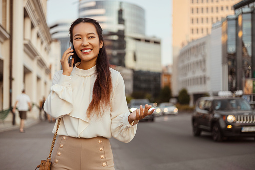 Young Asian Woman Standing in Big City Talking on Mobile Phone. Smiling Woman Making Business Call on Cell Walking on Busy Downtown Street Outdoors