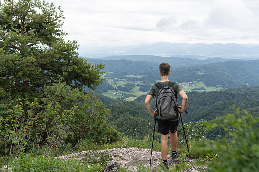 A teenage hiker with poles and a backpack while wandering to the top of Mount Nanos in Slovenia. At the top, a beautiful view of the valley. He sits and quenches his thirst with water from a water bottle.