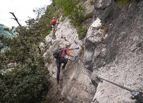 Woman climber going on via ferrata traveres. She is holding a safety harness. The climber has a purple T-shirt, a black backpack, a white helmet and a blue via ferrata set. Another climber in the background. Arco, Italy.