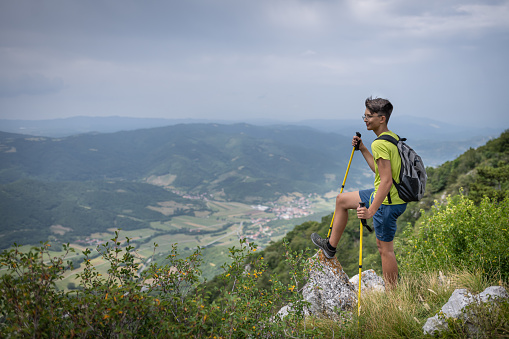A teenage hiker with poles and a backpack while wandering to the top of Mount Nanos in Slovenia. At the top, a beautiful view of the valley.  He stands triumphantly on the edge of the ridge and screams with raised hands.