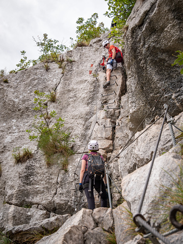 Climbers on via ferrata. One climber is standing on a ladder and talking to a woman standing at the bottom. Arco, Italy