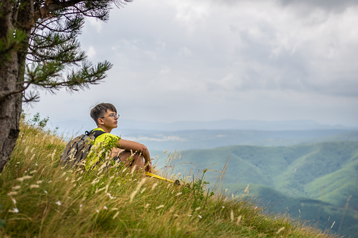 A teenage hiker with poles and a backpack while wandering to the top of Mount Nanos in Slovenia. At the top, a beautiful view of the valley. He sits triumphantly on the edge of the ridge and looks around, relaxing and enjoying the silence of the mountain.
