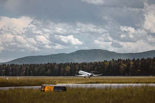 Full length side shot of a commercial airplane taking off on the airport runway surrounded by grass, trees and mountains on a day with a cloudy sky.