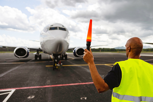 Waist-up side shot with blurred background of the back of a mid-adult bold Latin-American aircraft marshalling wearing a protective green and gray uniform and using one marshalling safety wand to do the 