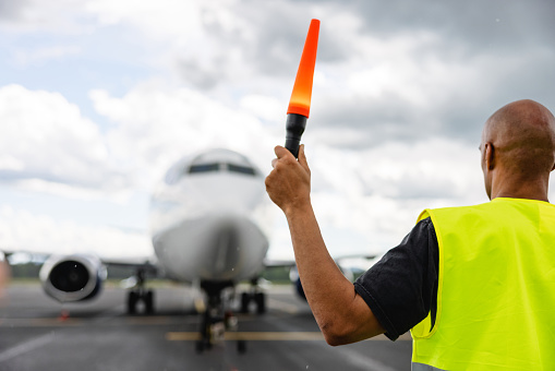 Waist-up side shot with blurred background of the back of a mid-adult bold Latin-American aircraft marshalling wearing a protective green and gray uniform and using one marshalling safety wand to do the 