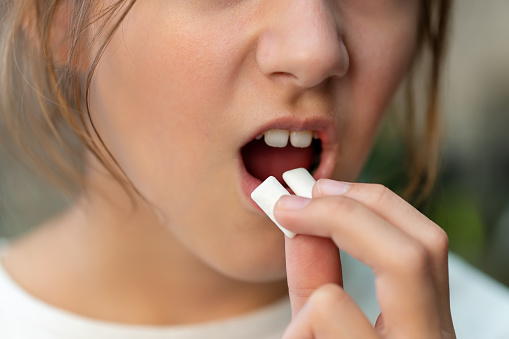 girl putting chewing gum piece into mouth on blurred background, closeup the hand puts the gum in the open mouth.