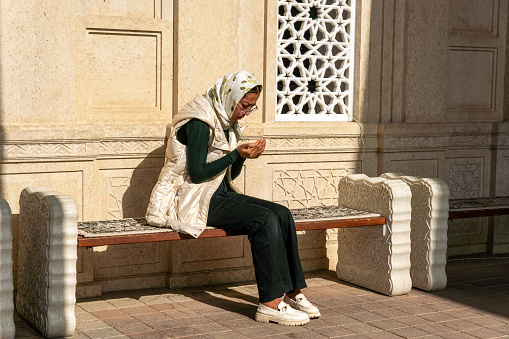 October 29, 2023. Samarkand, Uzbekistan: side view of beautiful asian muslim woman up palms open to light praying to allah. A Muslim girl is sitting on a bench in the courtyard of a mosque for women.