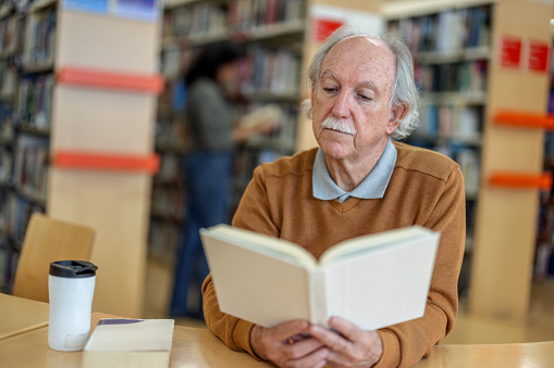 Waist-up side shot with blurred background of a gray hair senior Caucasian man seated reading a book at a public library while having a cup of coffee.