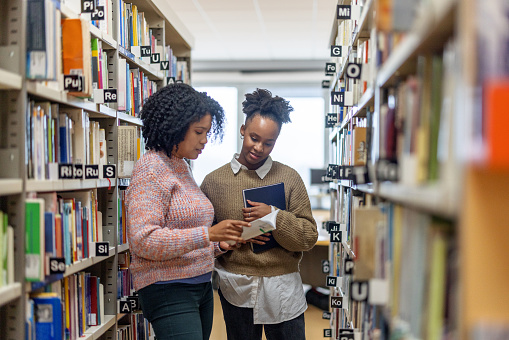 Three-quarter side shot with blurred background of a Latin-American female helping a younger African-American classmate to find a book to prepare herself for an important exam at the university library.