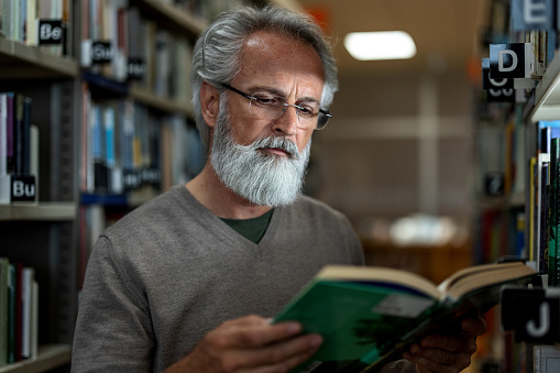 Close-up side shot with blurred background of a gray-haired senior Caucasian retired man reading a book of his interest at the city public library during the afternoon.