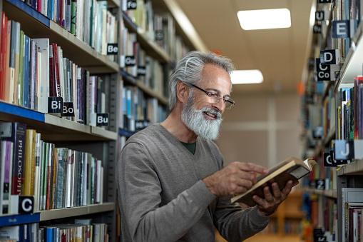Waist-up side shot of a gray-haired retired man reading a book of his interest while standing between to big bookshelves at the city library during the afternoon.