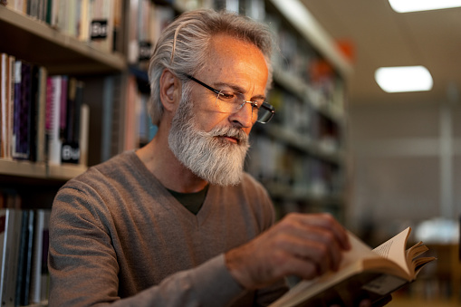 Waist-up side shot of a gray-haired retired man reading a book of his interest while standing between to big bookshelves at the city library during the afternoon.