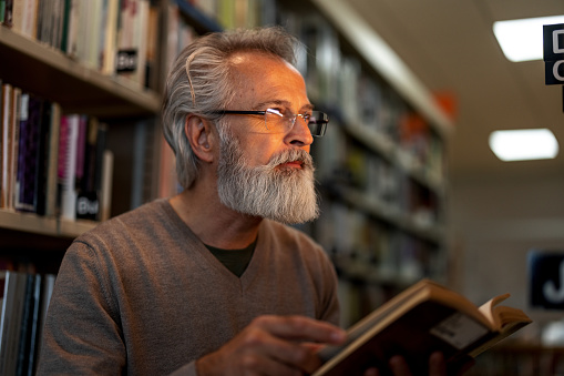 Waist-up side shot of a gray-haired retired man looking away while holding a book and standing between to big bookshelves at the city library during the afternoon.