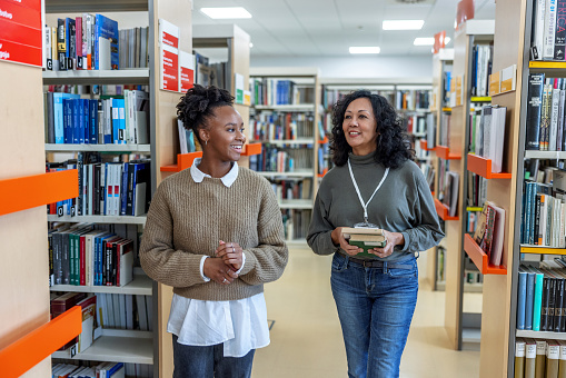 Three-quarter front shot with blurred background of a Latin-American female librarian helping to find a book to a young African-American student to prepare for an important exam while walking through the university library.