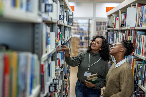 Aerial three-quarter front shot of a young African-American student being helped by a Latin-American female librarian to find some books for her final exam preparation at the university library.