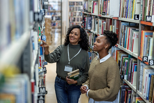 Aerial three-quarter front shot of a young African-American student being helped by a Latin-American female librarian to find some books for her final exam preparation at the university library.
