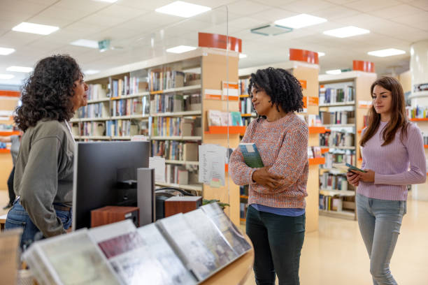 Three-quarter side shot with blurred background of a friendly and casually dressed adult Latin-American student checking out some books from the local college library, while being help by the female librarian.