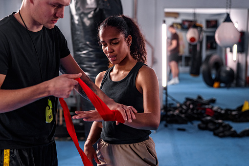 Athletic mid adult White male trainer helping a young Hispanic woman to wrap her hands before a boxing session. They are indoors in a boxing studio.