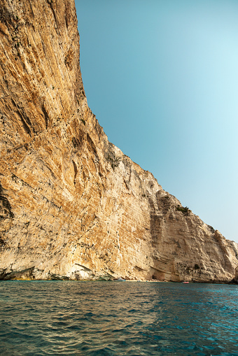 Majestic high wall of a cliff over the calm blue sea in Greece. The sun is shinning and illuminating the coastline.