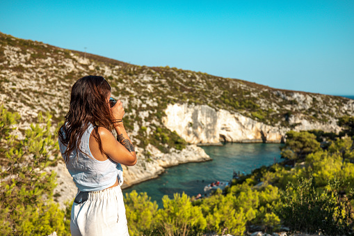 A young adult caucasian female tourist taking a photo of the amazing bay in  Greece. She is enjoying the view as she is standing on the top of the hill with a beautiful view of the nature. The sky is blue and the water is clear. The bay is surrounded by a stone beach and some high cliffs.