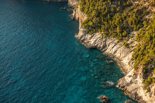 Aerial view of the beautiful clear blue water and the rocky shoreline in Greece. The cliffs over the blue sea are rocky and steep. Tops of the cliffs are covered in natural mediterranean vegetation. The sun is illuminating the coast.