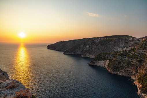 Beautiful view of the seascape in Greece during a sunset. The sun is going down and the sky is becoming orange. The sea is calm and surrounded by high steep cliffs.