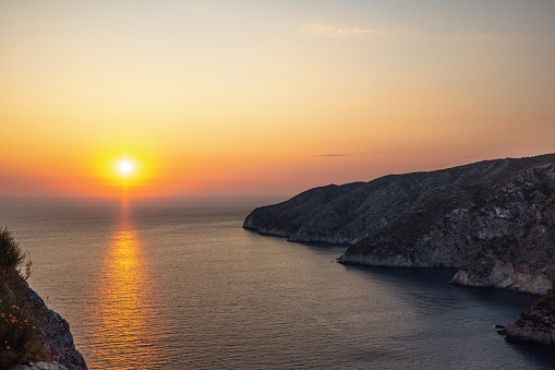 Amazing sunset from the top of the cliff in Greece. The sun is setting and the sky is orange. The coast and the cliffs are dark. There is a reflection of the sun in the calm sea surface.