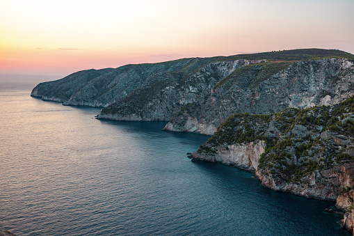 Beautiful shoreline in Greece during a sunset. The sky is orange and purple as the sun is setting. The cliffs are barely illuminated. The water is calm and blue.